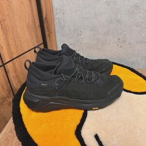 May include: A pair of black hiking boots with dark gray laces and a Vibram sole. The boots have a Gore-Tex label and are placed on a yellow and white rug with a black border. The background is a wooden surface and a gray wall.