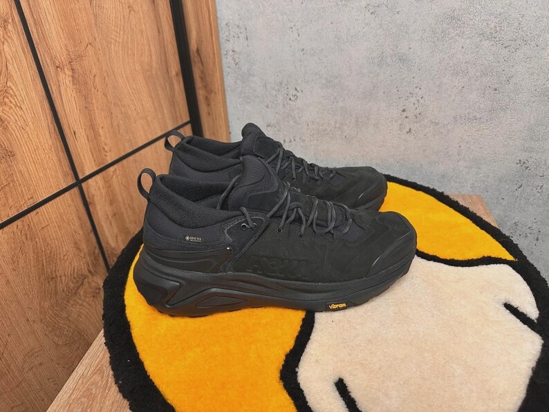 May include: A pair of black hiking boots with dark gray laces and a Vibram sole. The boots have a Gore-Tex label and are placed on a yellow and white rug with a black border. The background is a wooden surface and a gray wall.