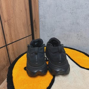 May include: A pair of black athletic shoes with thick soles, viewed from the back. The shoes have the text "KAHA 3" printed on the heel. They are placed on a yellow and white patterned rug.