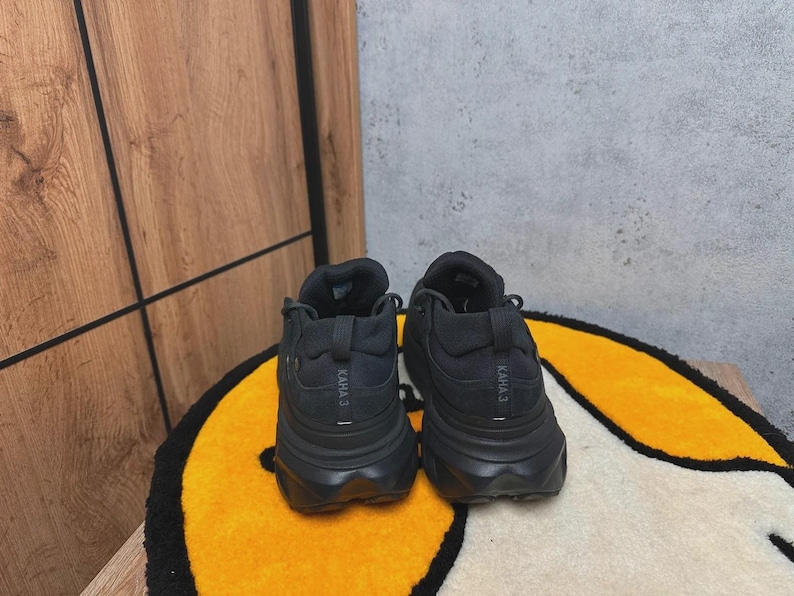 May include: A pair of black athletic shoes with thick soles, viewed from the back. The shoes have the text "KAHA 3" printed on the heel. They are placed on a yellow and white patterned rug.