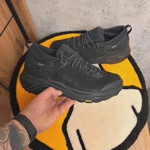 May include: Pair of black hiking boots with the brand name "HOKA" visible on the side. The boots have a rugged sole and are designed for outdoor activities. The boots are sitting on a yellow and white rug.