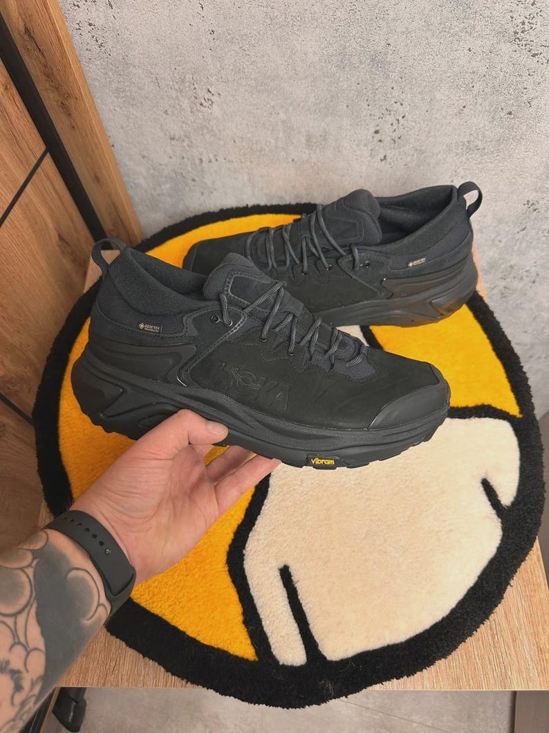 May include: Pair of black hiking boots with the brand name "HOKA" visible on the side. The boots have a rugged sole and are designed for outdoor activities. The boots are sitting on a yellow and white rug.