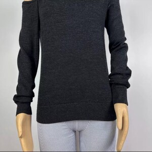 May include: Dark gray sweater with long sleeves and a cold shoulder design. The sweater has a crew neck and ribbed cuffs. The model is wearing striped pants.