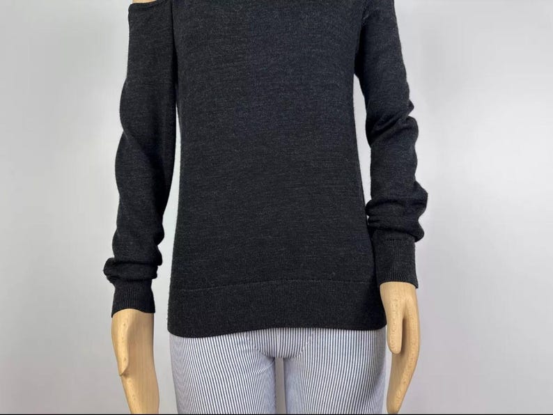 May include: Dark gray sweater with long sleeves and a cold shoulder design. The sweater has a crew neck and ribbed cuffs. The model is wearing striped pants.