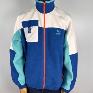 May include: A colour-blocked fleece jacket in blue, white, and teal. The jacket features a high collar, a full-length orange zip, and a white pocket on the chest. The Puma logo is visible on the chest. The jacket is displayed on a mannequin.
