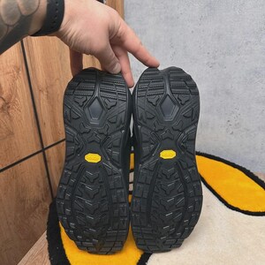 May include: A pair of black hiking boots, soles facing up, showcasing a rugged tread pattern. The soles have a yellow Vibram logo. The boots are on a yellow and white rug.