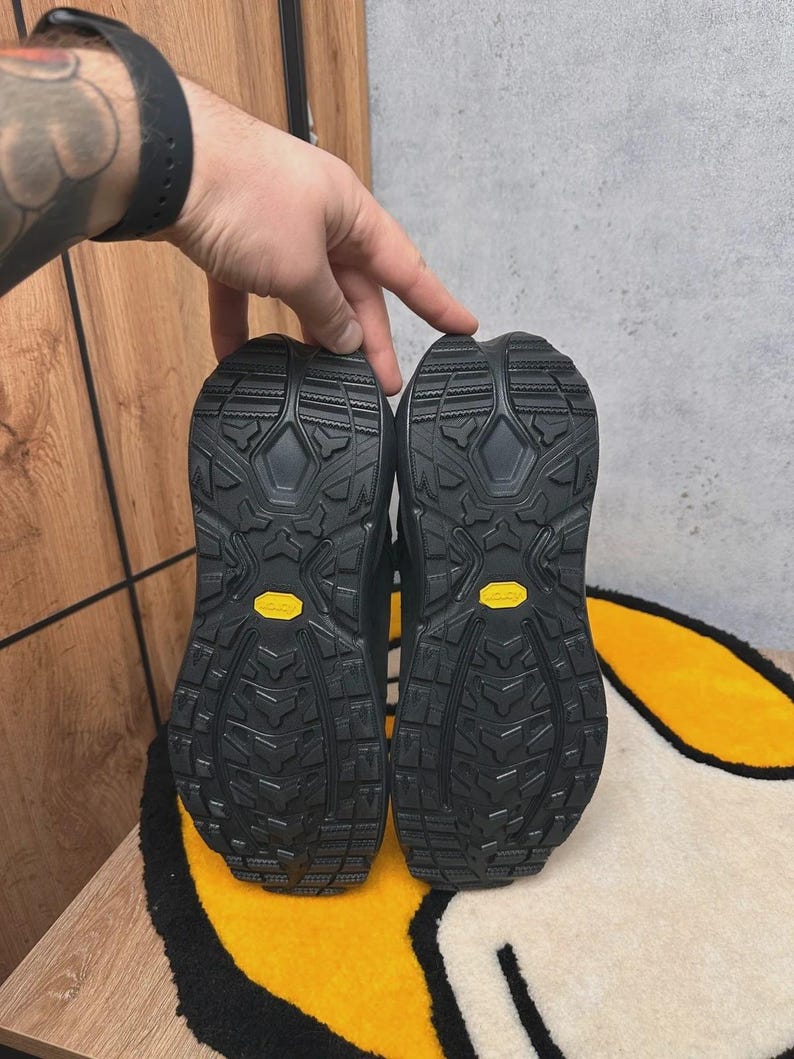 May include: A pair of black hiking boots, soles facing up, showcasing a rugged tread pattern. The soles have a yellow Vibram logo. The boots are on a yellow and white rug.