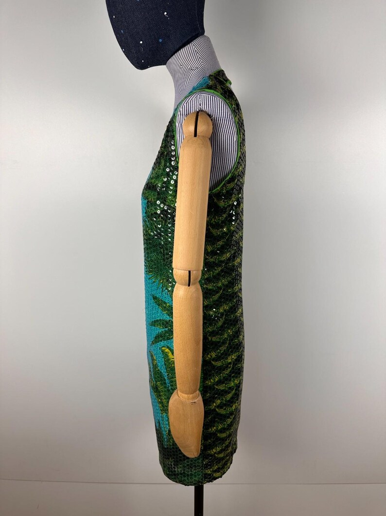 May include: A sleeveless dress with a tropical design. The dress features a vibrant pattern of green palm leaves and blue accents, embellished with sequins. The dress is displayed on a mannequin.