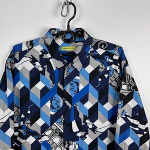 May include: A patterned button-down shirt in blue, grey, and white. The design features geometric shapes and a collared neckline. The label reads "VERSACE JEANS".