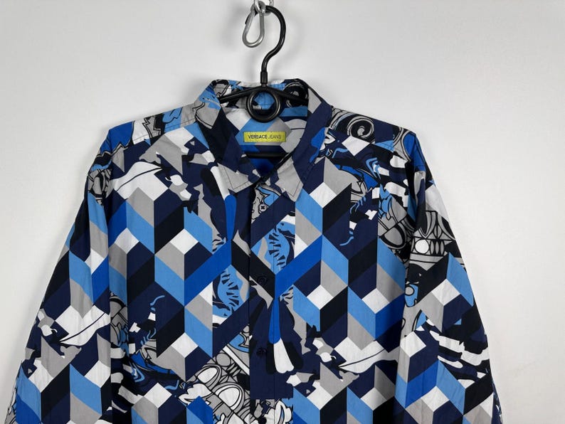 May include: A patterned button-down shirt in blue, grey, and white. The design features geometric shapes and a collared neckline. The label reads "VERSACE JEANS".
