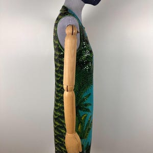 May include: A sleeveless dress with a vibrant tropical design. The dress features a green and black patterned top, transitioning to a turquoise blue base with a palm tree motif. The dress is displayed on a wooden mannequin.