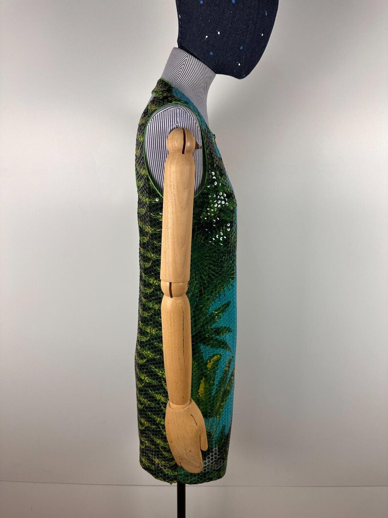 May include: A sleeveless dress with a vibrant tropical design. The dress features a green and black patterned top, transitioning to a turquoise blue base with a palm tree motif. The dress is displayed on a wooden mannequin.