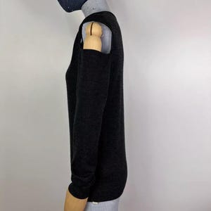 May include: A dark gray sweater with long sleeves and shoulder cutouts. The sweater is layered over a collared shirt with thin blue and white stripes. A dark blue beanie and a face mask are also visible.