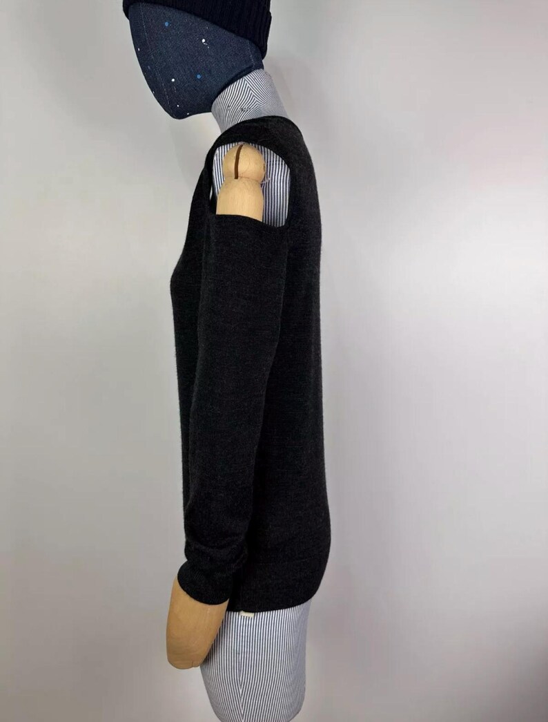May include: A dark gray sweater with long sleeves and shoulder cutouts. The sweater is layered over a collared shirt with thin blue and white stripes. A dark blue beanie and a face mask are also visible.
