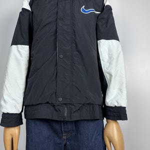May include: Black and white color block jacket with a blue Nike swoosh logo on the chest. The jacket has a snap button closure and a zipper.