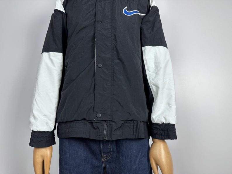 May include: Black and white color block jacket with a blue Nike swoosh logo on the chest. The jacket has a snap button closure and a zipper.