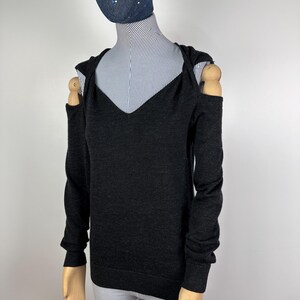 May include: A dark gray, long-sleeved sweater with a V-neck and open shoulders. The sweater is displayed on a mannequin with a striped shirt underneath. The mannequin also wears a dark blue beanie and a denim face covering with white polka dots.