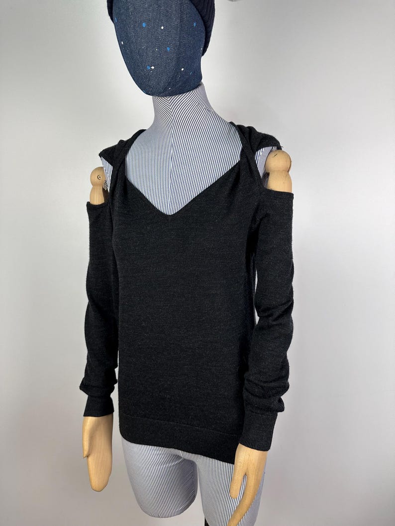 May include: A dark gray, long-sleeved sweater with a V-neck and open shoulders. The sweater is displayed on a mannequin with a striped shirt underneath. The mannequin also wears a dark blue beanie and a denim face covering with white polka dots.