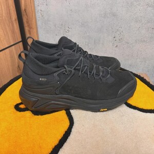May include: Pair of black hiking shoes with a thick sole, laces, and the word "HOKA" embossed on the side. The shoes are made of a suede-like material and have a "GORE-TEX" label. The soles have a "Vibram" logo.