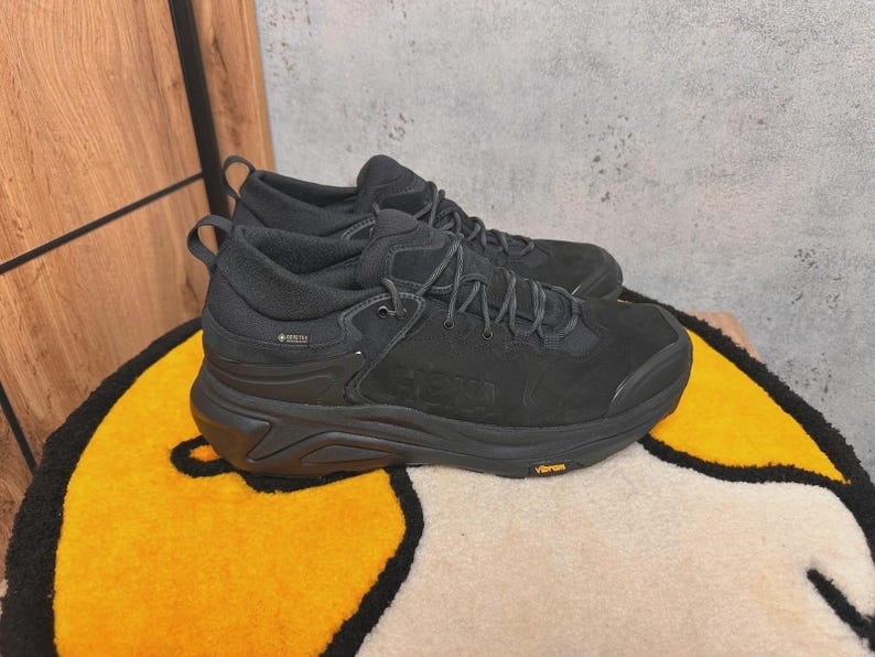 May include: Pair of black hiking shoes with a thick sole, laces, and the word "HOKA" embossed on the side. The shoes are made of a suede-like material and have a "GORE-TEX" label. The soles have a "Vibram" logo.
