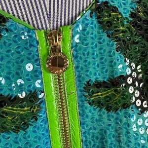 May include: Close-up of a teal sequined garment with a palm tree design. The garment features a bright green zipper and trim, and a gold zipper pull. The neckline is lined with a blue and white striped fabric.