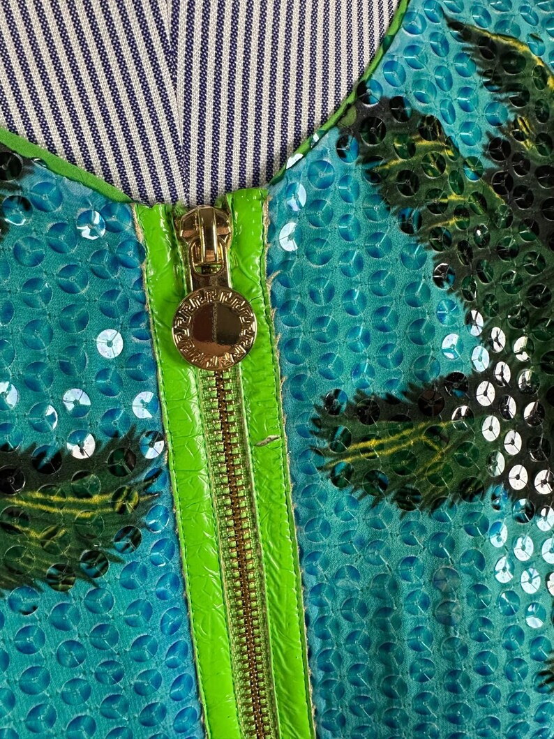 May include: Close-up of a teal sequined garment with a palm tree design. The garment features a bright green zipper and trim, and a gold zipper pull. The neckline is lined with a blue and white striped fabric.