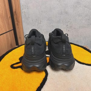 May include: A pair of black athletic shoes with thick soles, viewed from the back. The shoes have the text "KAHA 3" printed on the heel. They are placed on a yellow and black patterned rug.