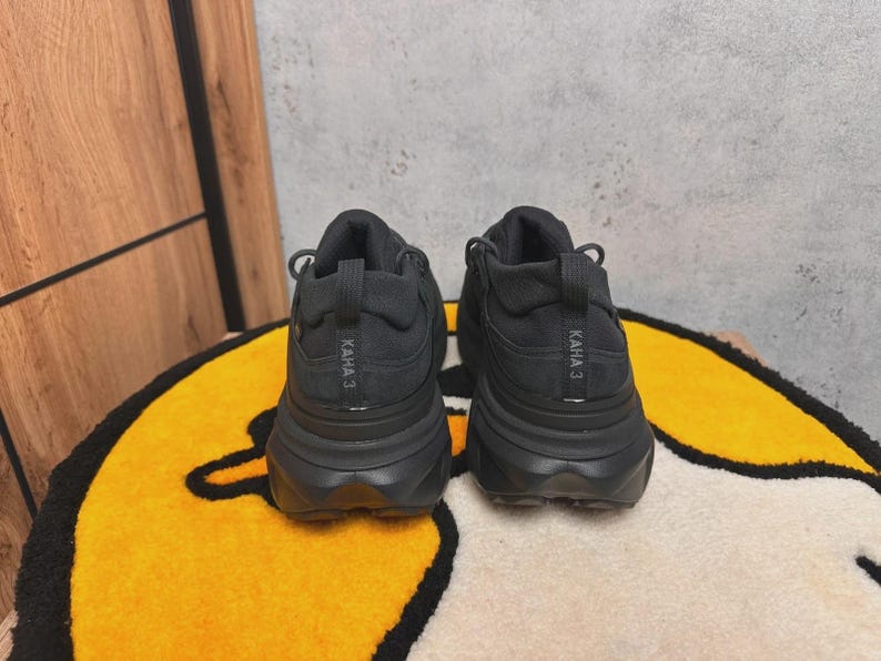 May include: A pair of black athletic shoes with thick soles, viewed from the back. The shoes have the text "KAHA 3" printed on the heel. They are placed on a yellow and black patterned rug.