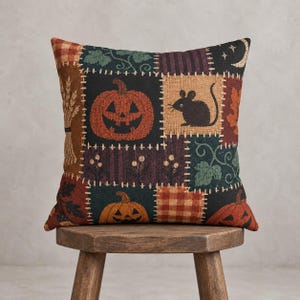 May include: A square throw cushion featuring a patchwork design with autumn-themed motifs including orange jack-o'-lanterns, a black mouse silhouette, wheat stalks, green leaves, and a crescent moon on a dark background with plaid and striped patterns.