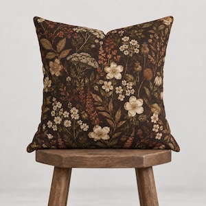 May include: A square throw pillow featuring a dark brown background with a dense, repeating pattern of various wildflowers, leaves, and seed pods in shades of cream, beige, muted orange, and deep green, creating a detailed botanical print.