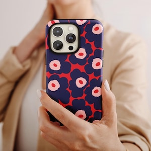 Puede incluir: Una funda de teléfono azul marino con un fondo rojo y un estampado floral de grandes flores azules estilizadas con centros rosas. La funda está sujeta en la mano de una persona.