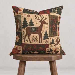 Peut inclure: Un coussin carré au motif patchwork représentant un cerf brun central, des sapins, des pommes de pin, des baies rouges et des lapins. Le tissu présente une texture matelassée avec une palette de couleurs crème, vert forêt et rouge foncé.