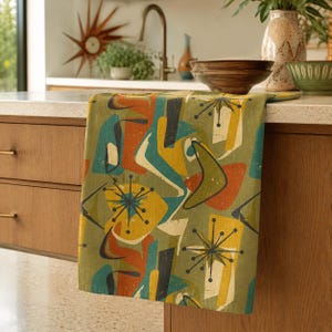 May include: A kitchen towel featuring a repeating pattern of abstract shapes in olive green, mustard yellow, burnt orange, and teal. The design includes starburst motifs and boomerang shapes printed on a textured fabric with a mid-century aesthetic.