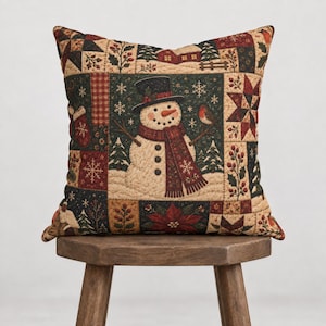 Peut inclure: Un coussin carré au motif patchwork représentant un bonhomme de neige central portant un haut-de-forme noir et une écharpe rouge. Le tissu présente des motifs de fêtes incluant des poinsettias rouges, des étoiles, du houx et des sapins.