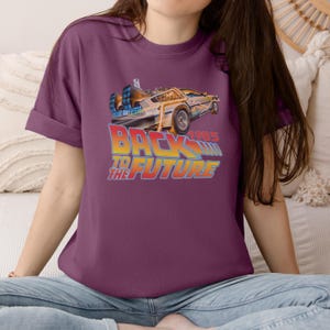 May include: Plum-colored t-shirt featuring a graphic of the DeLorean time machine from the movie "Back to the Future." The text "BACK TO THE FUTURE" is in a gradient of yellow to orange, with "1985" above the text.