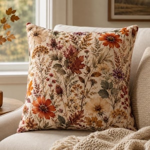 Op de afbeelding: Een vierkant sierkussen met een crèmekleurige achtergrond en een gedetailleerde botanische print. Het ontwerp toont diverse herfstbloemen, gedroogde grassen en herfstbladeren in tinten van roestoranje, dieprood, bruin en gedempt paars.