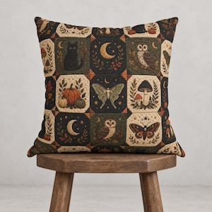 Peut inclure: Un coussin carré au motif patchwork illustrant des chats noirs, des hiboux, des papillons de nuit, des papillons, des citrouilles, des champignons et des croissants de lune dans des tons noir, crème, orange et vert sur un tissu matelassé.