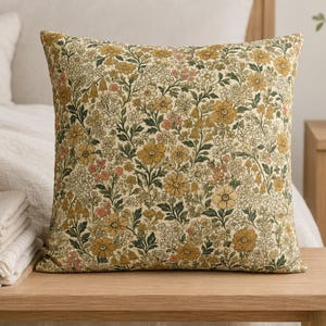 May include: A square throw pillow featuring a dense, intricate botanical print with mustard yellow, muted orange, and dark green floral patterns set against a cream-colored background. The design covers the entire front surface of the pillow.
