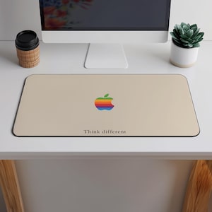 Puede incluir: Alfombrilla de ratón rectangular beige con borde negro. Presenta un logotipo de Apple colorido y la frase "Think different" en texto negro. Un monitor de ordenador, una pequeña planta y una taza de café están en el fondo.