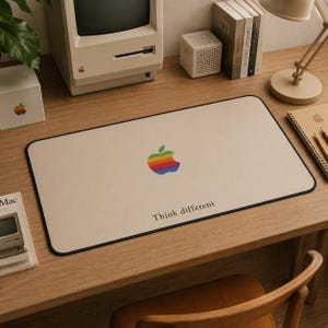 Puede incluir: Una alfombrilla de escritorio rectangular con superficie beige y borde negro. El centro presenta un logotipo de manzana con los colores del arcoíris sobre el texto negro "Think different". La alfombrilla está sobre un escritorio de madera.