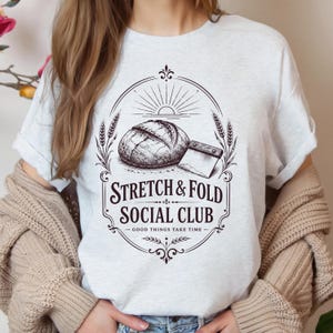 Può includere: T-shirt grigio chiaro con una grafica raffigurante una pagnotta, un coltello da pane e la scritta "STRETCH & FOLD SOCIAL CLUB - GOOD THINGS TAKE TIME". Il design include spighe di grano e un sole.