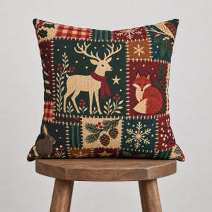 May include: A square throw pillow featuring a patchwork design with forest animals including a deer in a red scarf, a fox, and a rabbit. The pattern includes red and green plaid, pine cones, holly berries, and white snowflakes on a dark background.