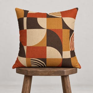 May include: A square throw pillow featuring a geometric pattern of semicircles, rectangles, and curved lines in shades of burnt orange, mustard yellow, dark brown, and cream. The fabric has a visible woven texture throughout the entire surface.