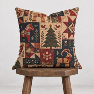Puede incluir: Un cojín cuadrado con un diseño de patchwork con temática navideña. La tela muestra ilustraciones de un caballo de Dala rojo, un árbol de Navidad decorado, una cabra beige, estrellas, copos de nieve, corazones y motivos florales en rojo, verde y beige.