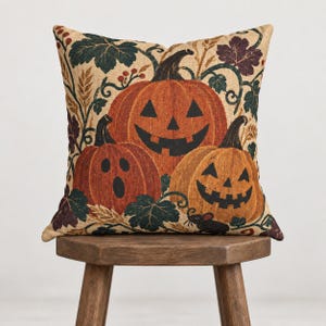 Peut inclure: Un coussin carré avec un fond en tissu beige imprimé de trois citrouilles orange en forme de jack-o'-lantern. Les citrouilles sont entourées de feuilles d'automne vert foncé et violettes, d'épis de blé et d'une petite souris noire en bas.
