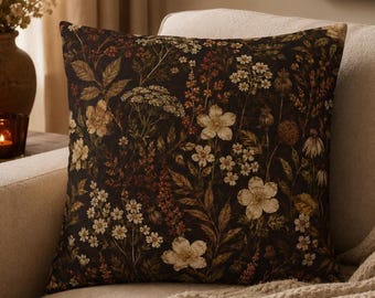 Op de afbeelding: Een vierkant sierkussen met een donkerbruine achtergrond en een dichte, gedetailleerde botanische print. Het patroon toont diverse wilde bloemen, bladeren en zaaddozen in tinten crème, gedempt goud, roest en donkergroen op de stof.