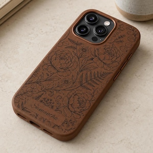 Puede incluir: Una funda de teléfono marrón con un patrón floral grabado en negro de peonías, helechos y hojas. La funda incluye una placa rectangular en la parte inferior con el nombre Samantha grabado en letra cursiva y un recorte para triple cámara.