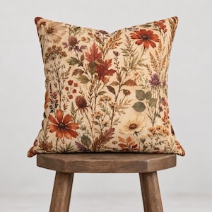 May include: A square throw pillow featuring a botanical print on a cream background. The pattern displays various wildflowers, autumn leaves, and grasses in shades of rust orange, deep red, muted yellow, brown, and sage green throughout the fabric.