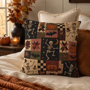 Op de afbeelding: Een vierkant sierkussen met een patchworkontwerp met Halloween-motieven, waaronder dansende skeletten, oranje pompoenen, zwarte vleermuizen, rode rozen en geometrische sterpatronen in een kleurenpalet van zwart, oranje, crème en donkerrood.