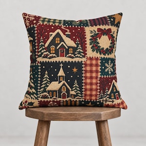 Könnte beinhalten: Ein quadratisches Zierkissen mit Patchwork-Design, das winterliche Szenen zeigt, darunter ein verschneites Haus, eine Kirche, einen Weihnachtskranz und Tannenbäume. Die Farbpalette umfasst Dunkelrot, Waldgrün und Creme mit karierten Akzenten.